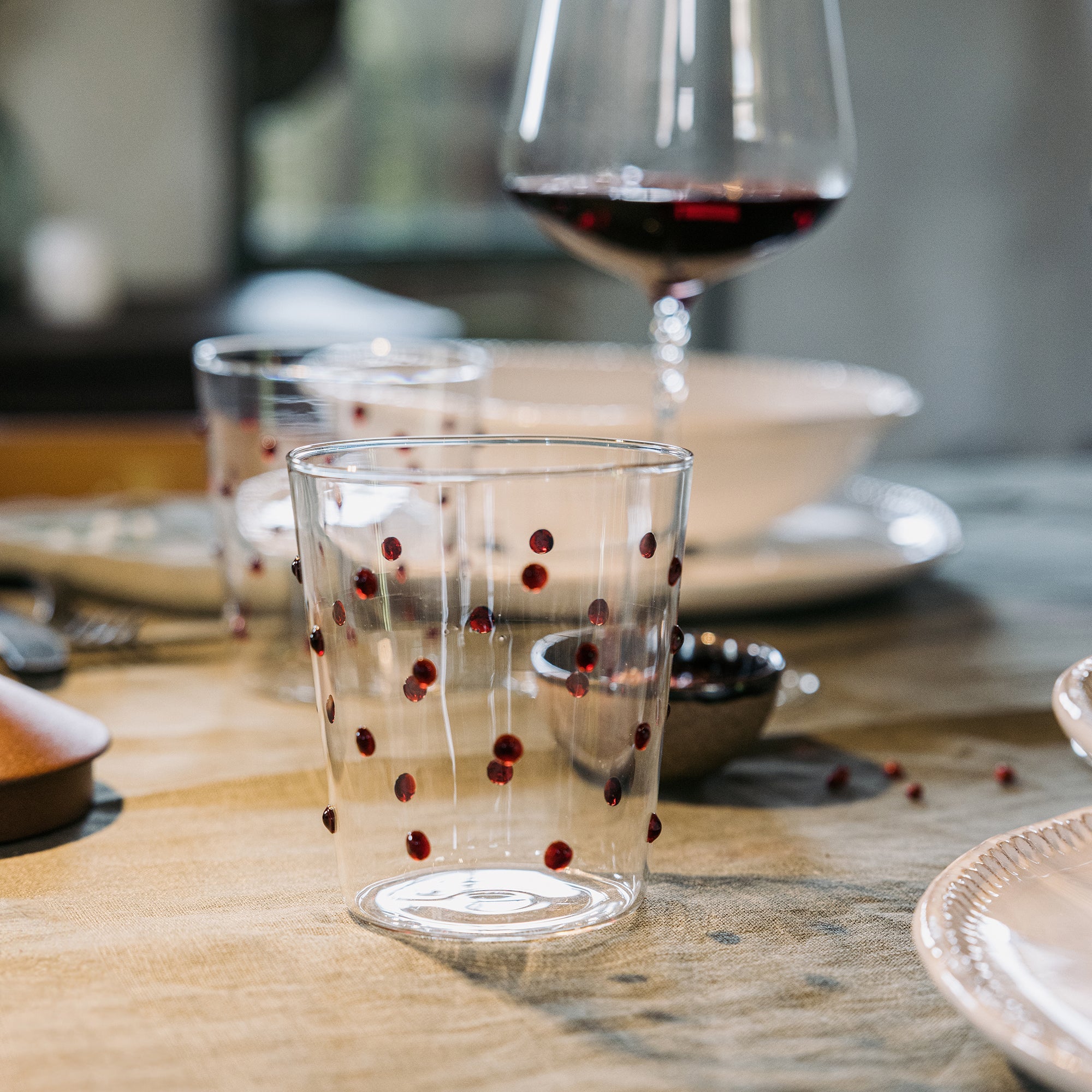 Bicchiere trasparente in vetro con pois rossi appoggiato su tavola imbandita con piatti beige e calice di vino rosso sullo sfondo.