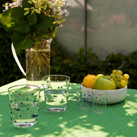 tavolo verde con due bicchieri in vetro trasparente con bolle colorate contenenti acqua, due bicchierini piccoli, vaso trasparente con fiori e