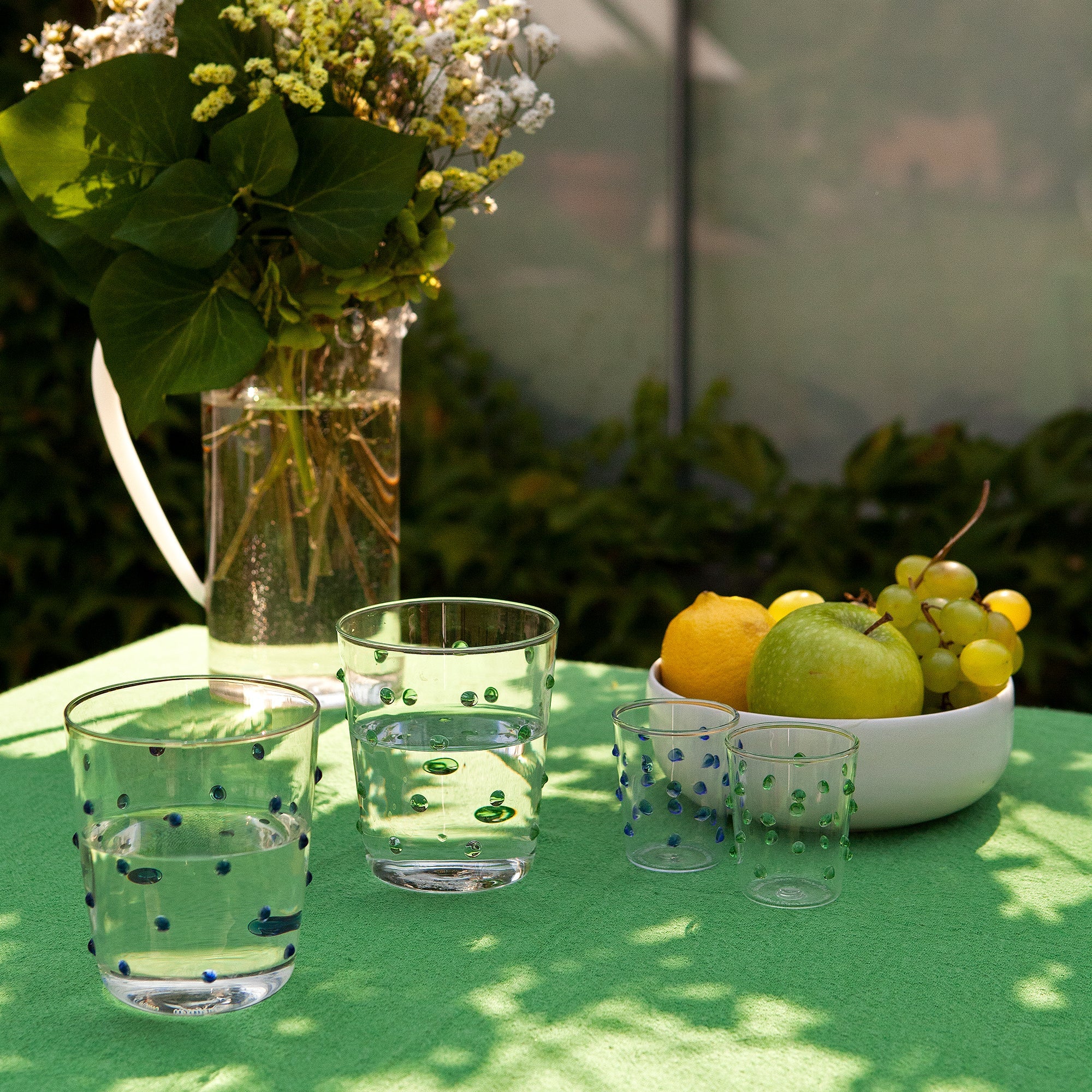 tavolo verde con due bicchieri in vetro trasparente con bolle colorate contenenti acqua, due bicchierini piccoli, vaso trasparente con fiori e