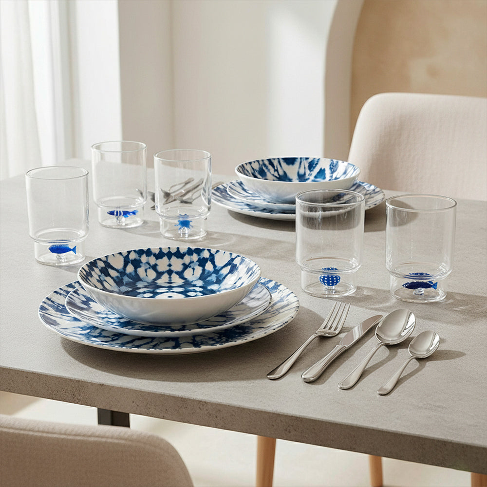 Set di stoviglie e bicchieri su tavolo grigio con piatti e ciotole bianchi con motivo blu marmorizzato e bicchieri trasparenti con decorazioni marine
