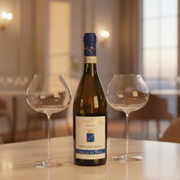 Bottiglia di vino bianco Benito Ferrara Greco di Tufo con tappo blu e due bicchieri di vetro trasparente su tavolo in ambiente elegante interno.