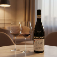 Bottiglia di vino rosso Borgogno Barolo 2018 e due calici con vino su tavolo, sfondo interno con lampada e poltrona beige.