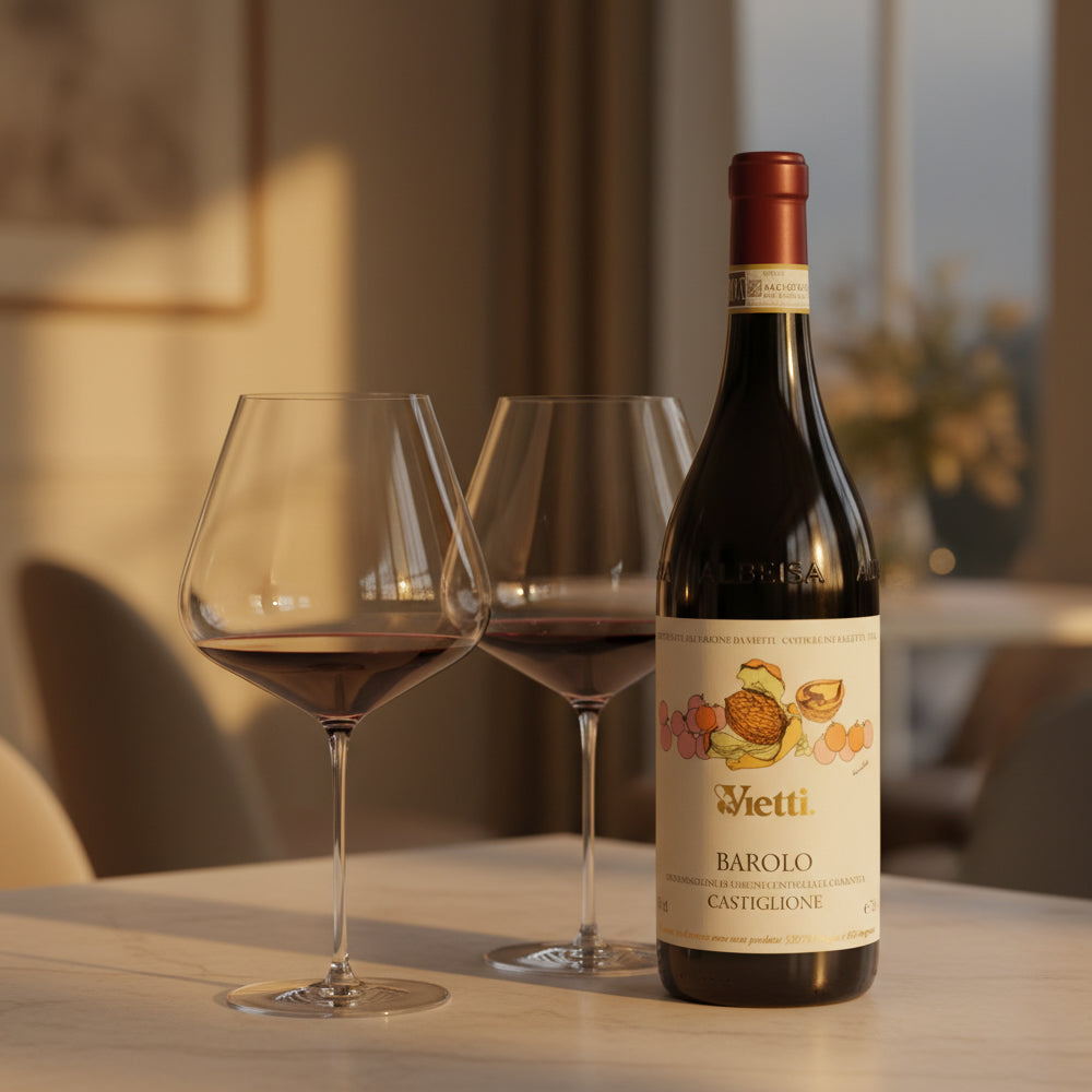 Bottiglia di vino rosso Vietti Barolo Castiglione con etichetta beige e disegni colorati, accompagnata da due bicchieri di vino su tavolo chiaro.