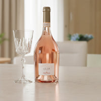 Bottiglia di vino rosato Alìe Frescobaldi in vetro trasparente e bicchiere di cristallo con gambo decorato su tavolo chiaro in interno luminoso.