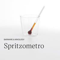 Bicchiere trasparente in vetro con bacchetta dosatrice con estremità tonda marrone e arancione su fondo bianco.