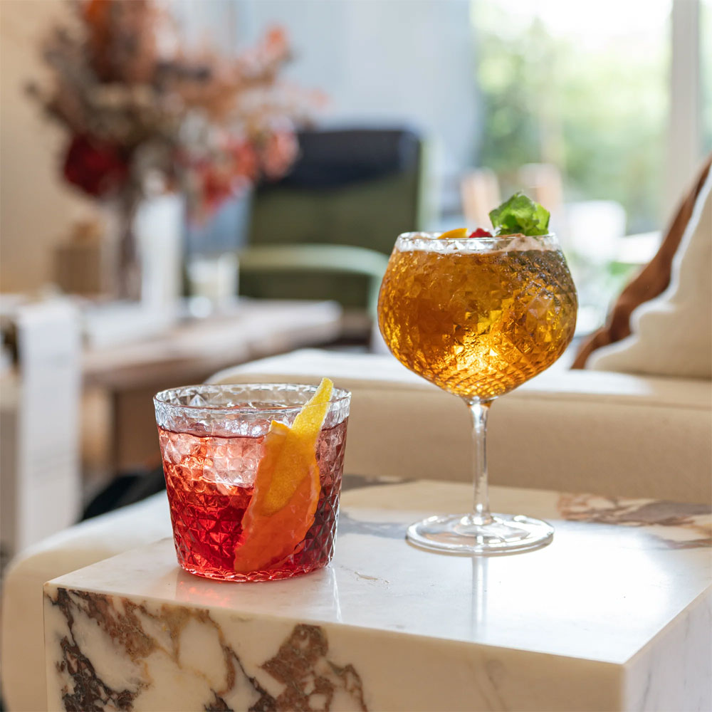 due cocktail in bicchieri di vetro sfaccettato, uno rosso con scorza di agrume e uno ambra con foglia di menta, su tavolo di marmo bianco