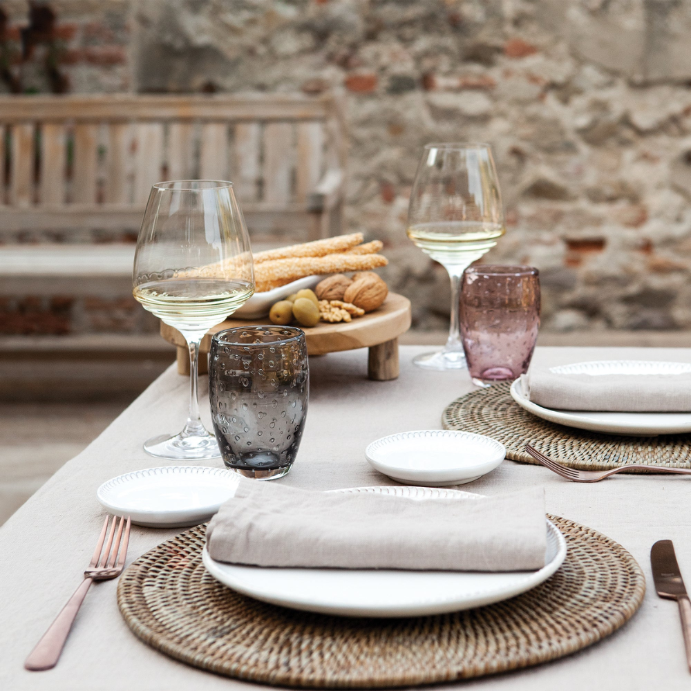 Tavola apparecchiata con piatti bianchi, tovaglioli beige, calici di vino bianco e bicchieri decorati su tovaglia chiara davanti a muro in