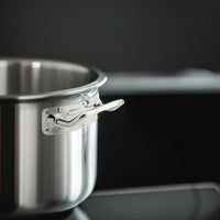 Pentola in acciaio inox lucido con manico corto, posizionata su piano cottura nero in ambiente cucina scura.