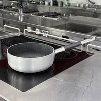 Pentola bassa rotonda in alluminio spazzolato con interno antiaderente nero e manico lungo in metallo con impugnatura nera, su piano