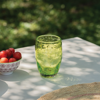 Bicchiere verde trasparente con decorazioni a bolle su tavolo bianco all'aperto, accanto a ciotola con prugne rosse.